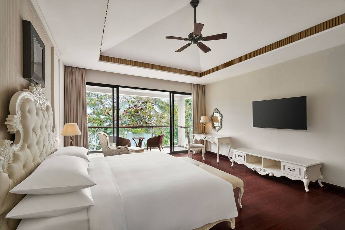 Een ruime slaapkamer met een groot bed, een plafondventilator, een televisie en een balkon met uitzicht op bomen.