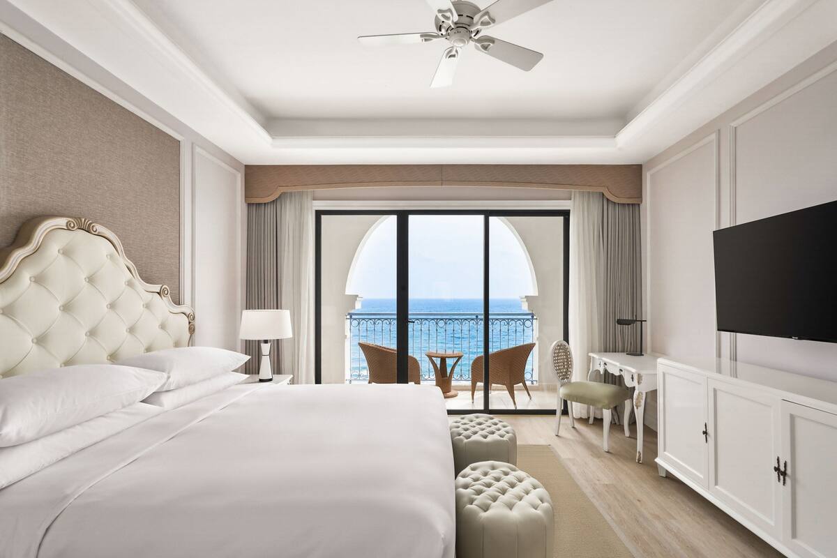 Hotelkamer met een groot bed, een plafondventilator, een televisie, een balkon met uitzicht op zee en een bureau met stoel.