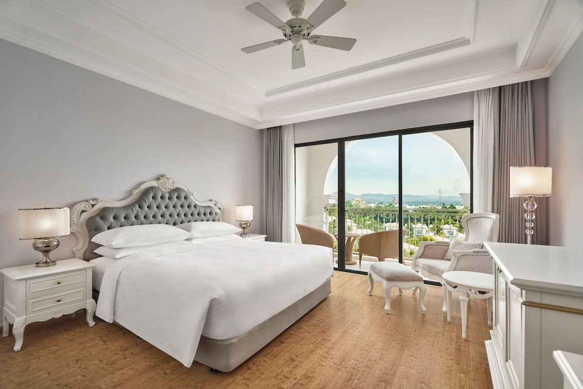 Ruime slaapkamer met een groot bed, een plafondventilator, een balkon met uitzicht en een zithoek met een tafel en stoelen.