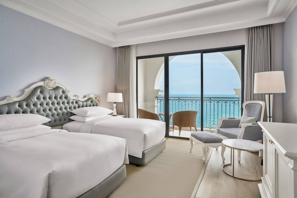 Een hotelkamer met twee bedden, een balkon met uitzicht op zee, en een zithoek met een tafel en stoelen.