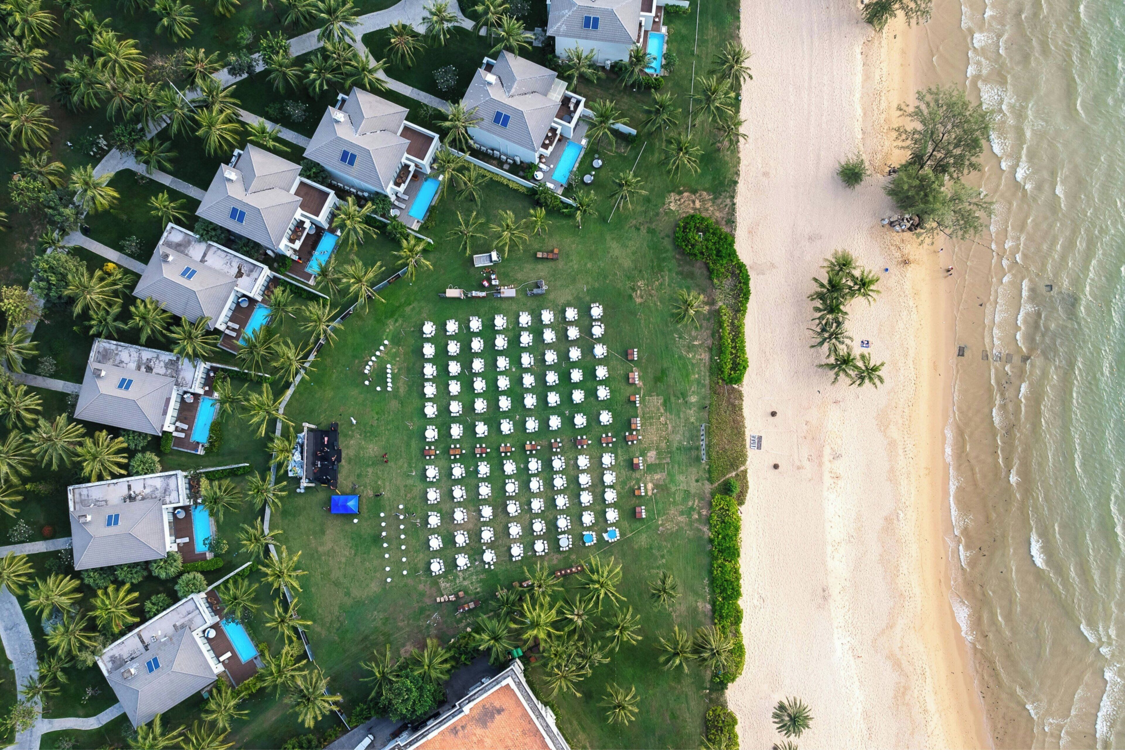 An aerial view of a resort with multiple bungalow-style buildings, a large lawn area with numerous white lounge chairs, and a sandy beach adjacent to the ocean.