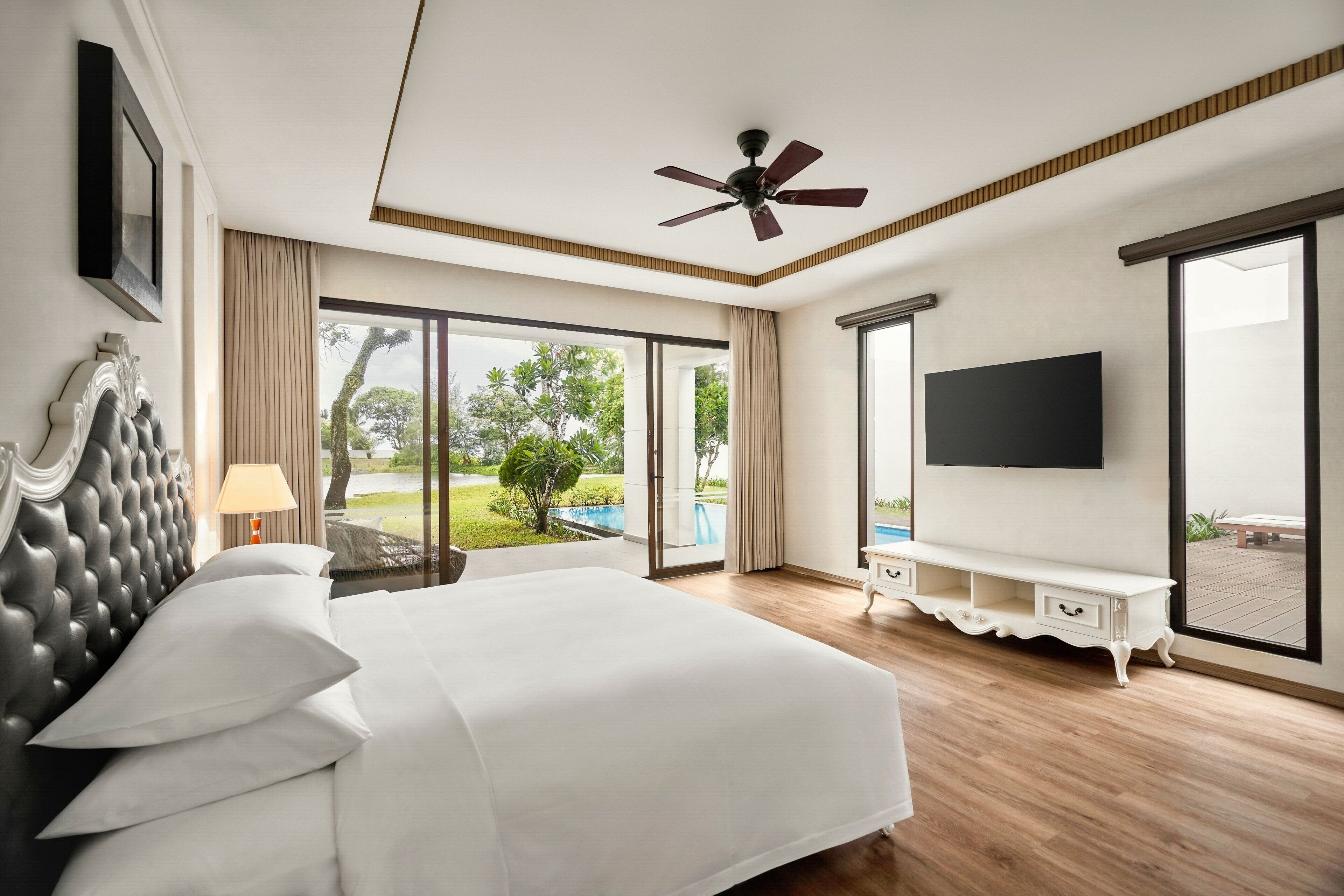 A hotel room with a large bed, a ceiling fan, a TV, and a view of a pool and greenery.