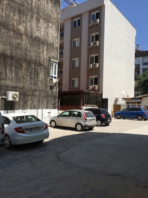 Ein Parkplatz mit mehreren Autos, ein mehrgeschossiges Gebäude mit Klimaanlagen und eine Betonmauer.