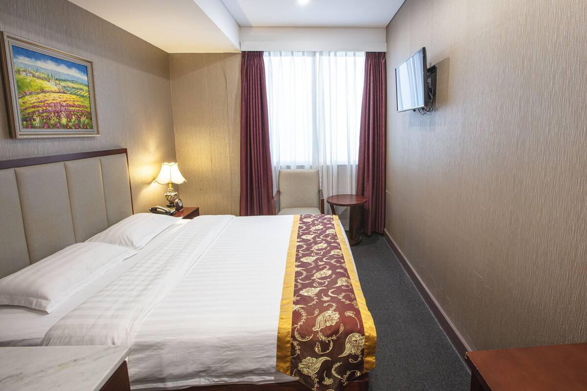 A hotel room with a bed, a nightstand, a lamp, a painting on the wall, and a window with curtains.