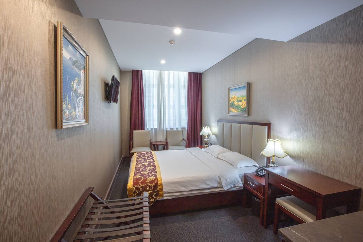 A hotel room with a large bed, bedside tables, a desk, a chair, a bench, a TV, and a painting on the wall.