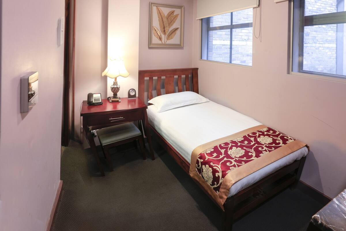 A small, neatly arranged bedroom with a single bed, a bedside table with a lamp, a chair, and a wall-mounted clock.