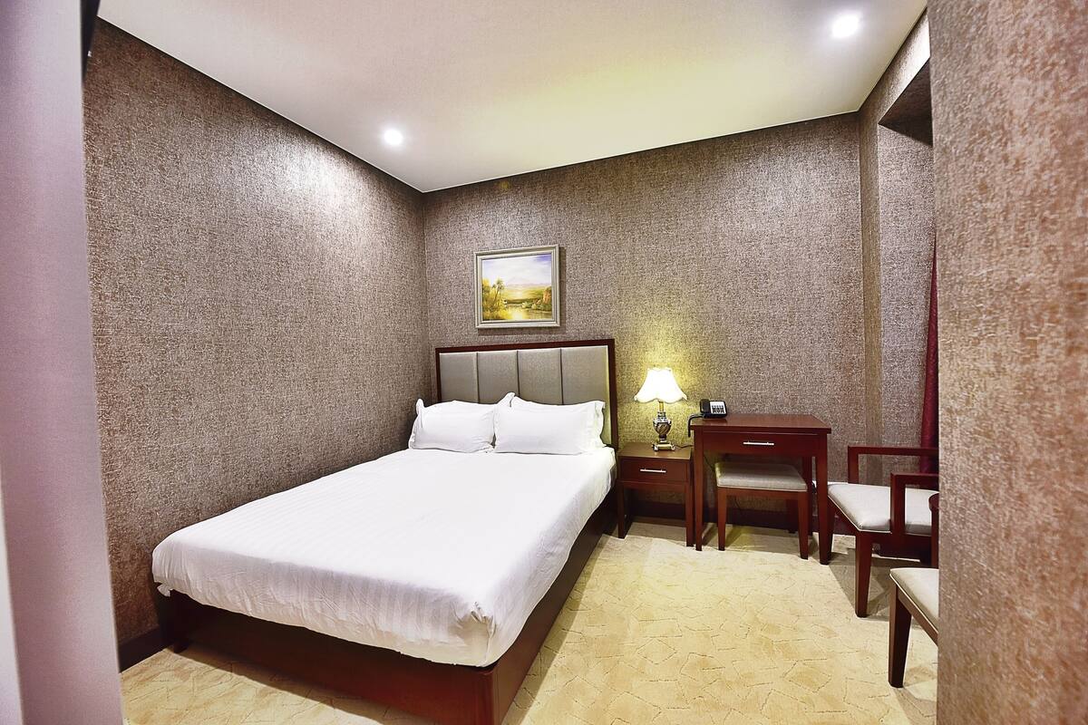 A hotel room with a bed, a desk with a chair, a lamp, and a painting on the wall.