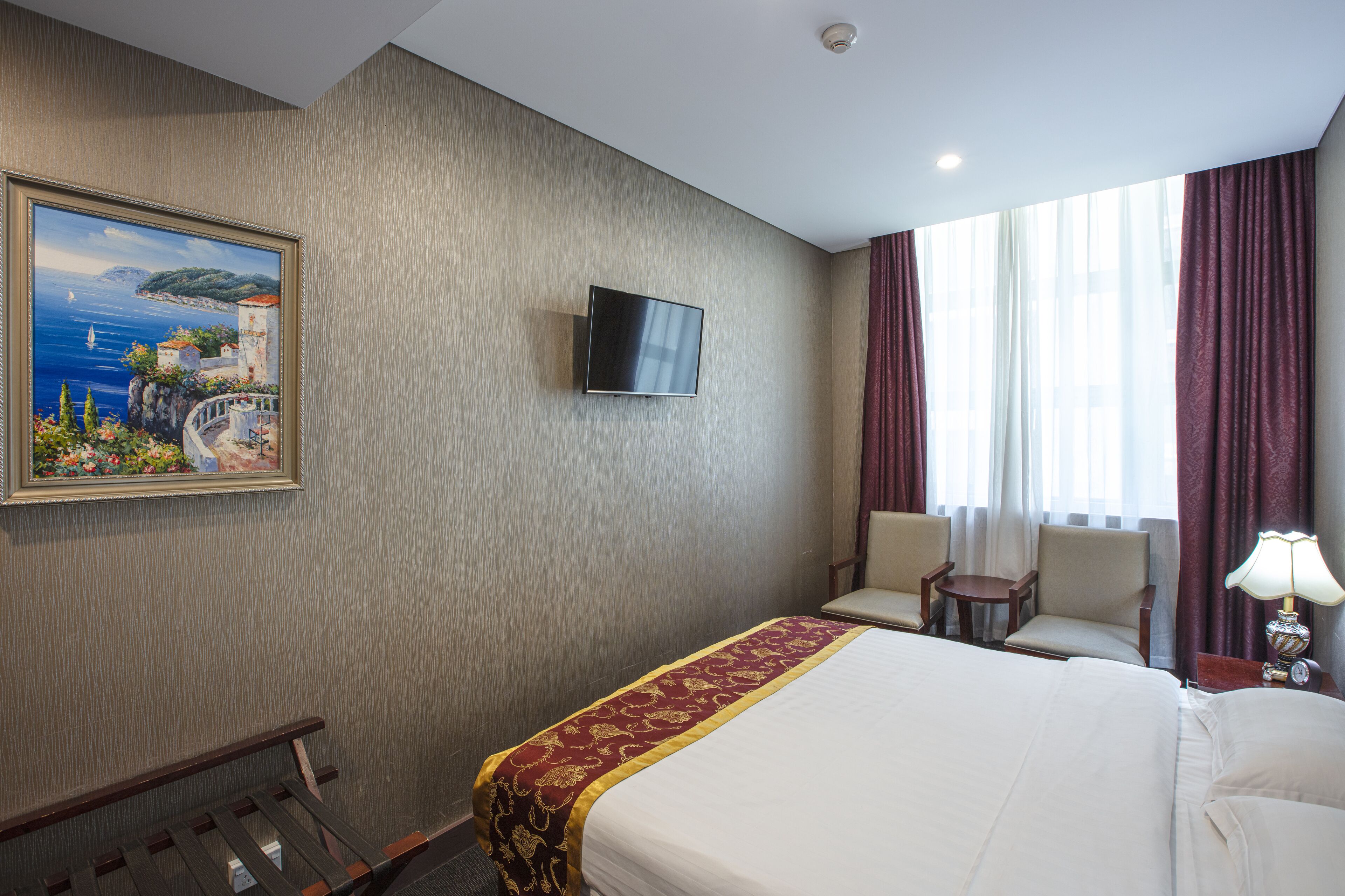 A hotel room with a large bed, a television mounted on the wall, a painting of a coastal scene, and a window with curtains.