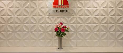A reception area with a decorative wall, a sign for the Megaboom City Hotel, and a vase with flowers.