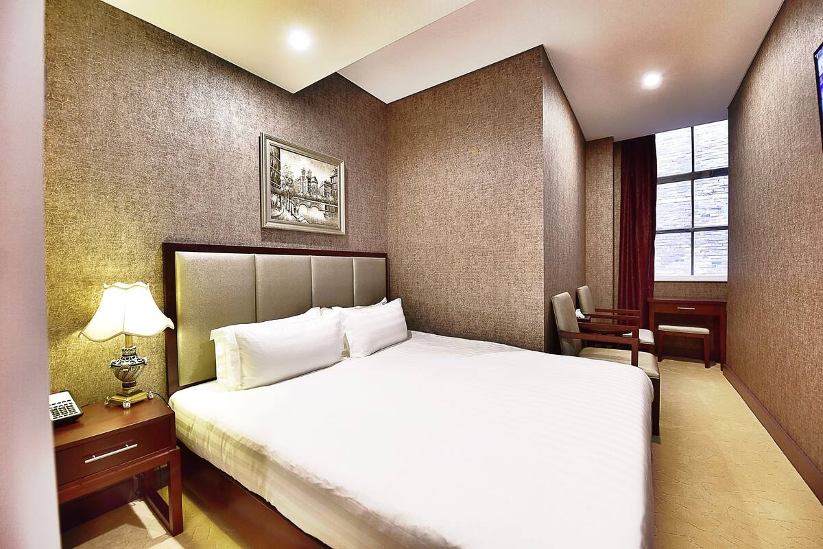 A hotel room with a large bed, a nightstand with a lamp, a desk, and a chair.