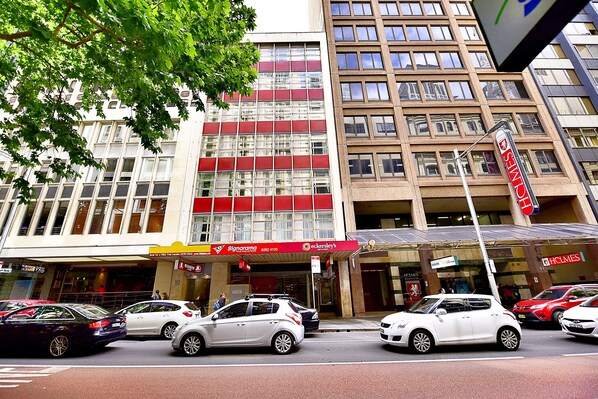 A street view with cars parked in front of a building with multiple shops, including a visible sign for 'Sign of the Times'. - Megaboom City Hotel (Sydney)