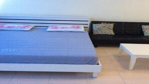 A bed with a grey mattress and white headboard, a black sofa with floral cushions, and a small white table. - Don Mueang-Challenger Impact Muangthong Thani (Pak Kret)