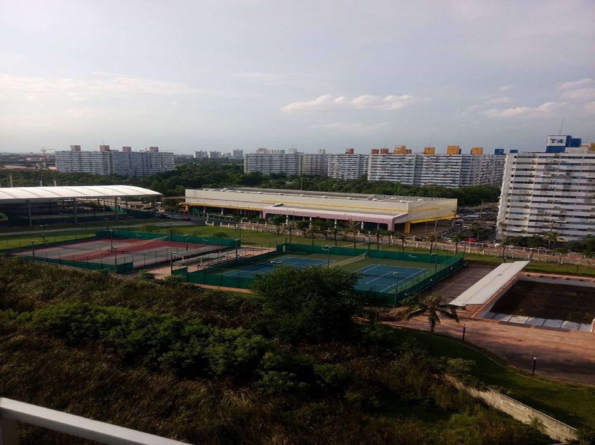 A sports complex with multiple tennis courts, a covered seating area, and a large building in the background.
