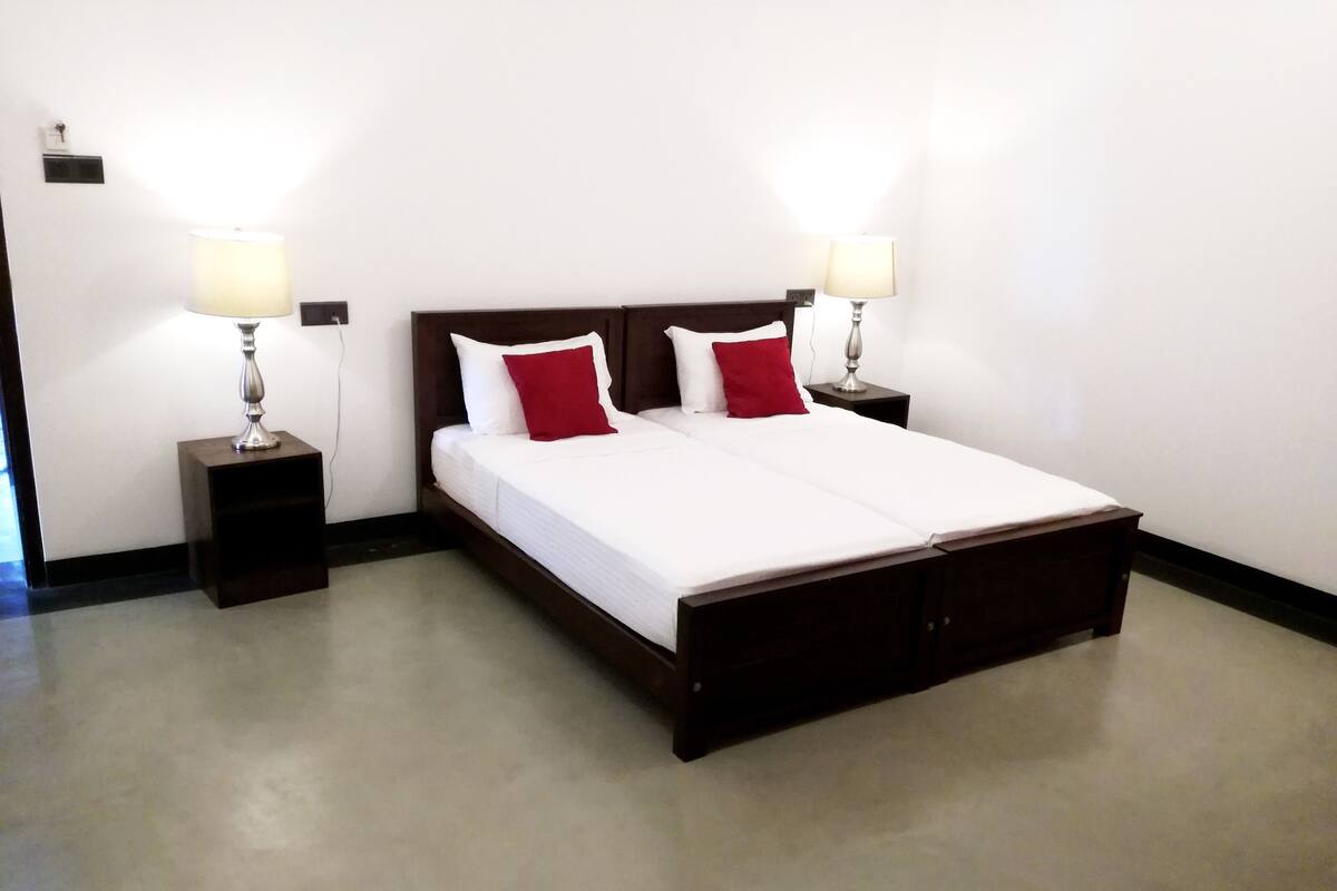 A neatly made double bed with white and red pillows, flanked by two bedside tables with lamps, in a room with plain white walls and a dark floor.