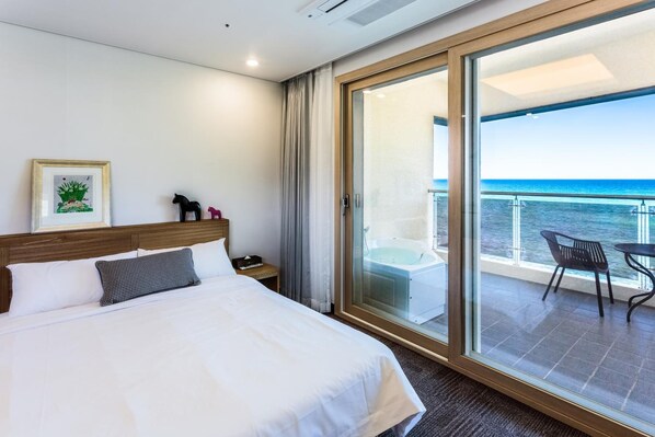 A hotel room with a large bed, a view of the ocean through a sliding glass door, and a balcony with a table and chairs.
