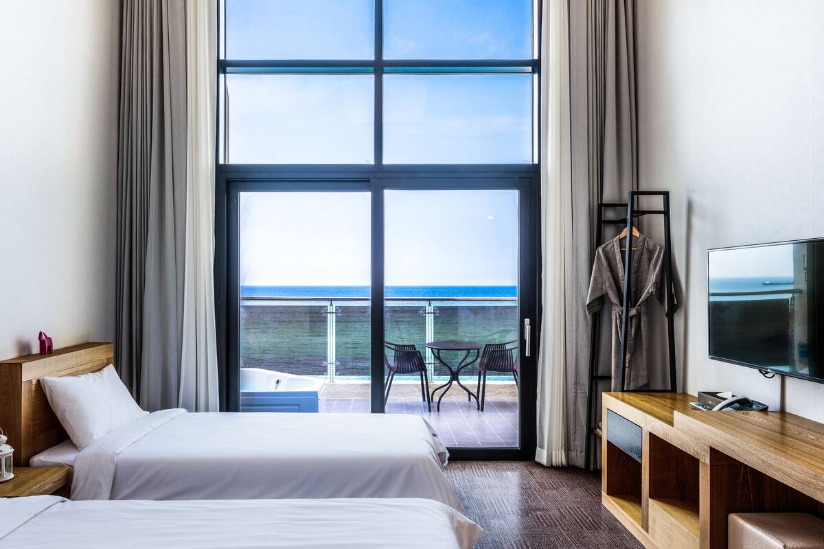 A hotel room with a large sliding glass door leading to a balcony with a view of the sea. There is a bed, a desk with a TV, and a chair.