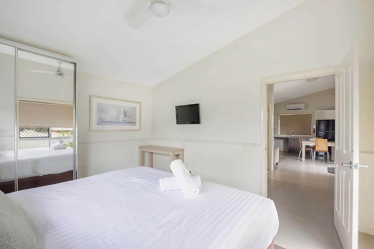 A hotel room with a bed, a bedside table, a TV, and a view of the kitchen and dining area.
