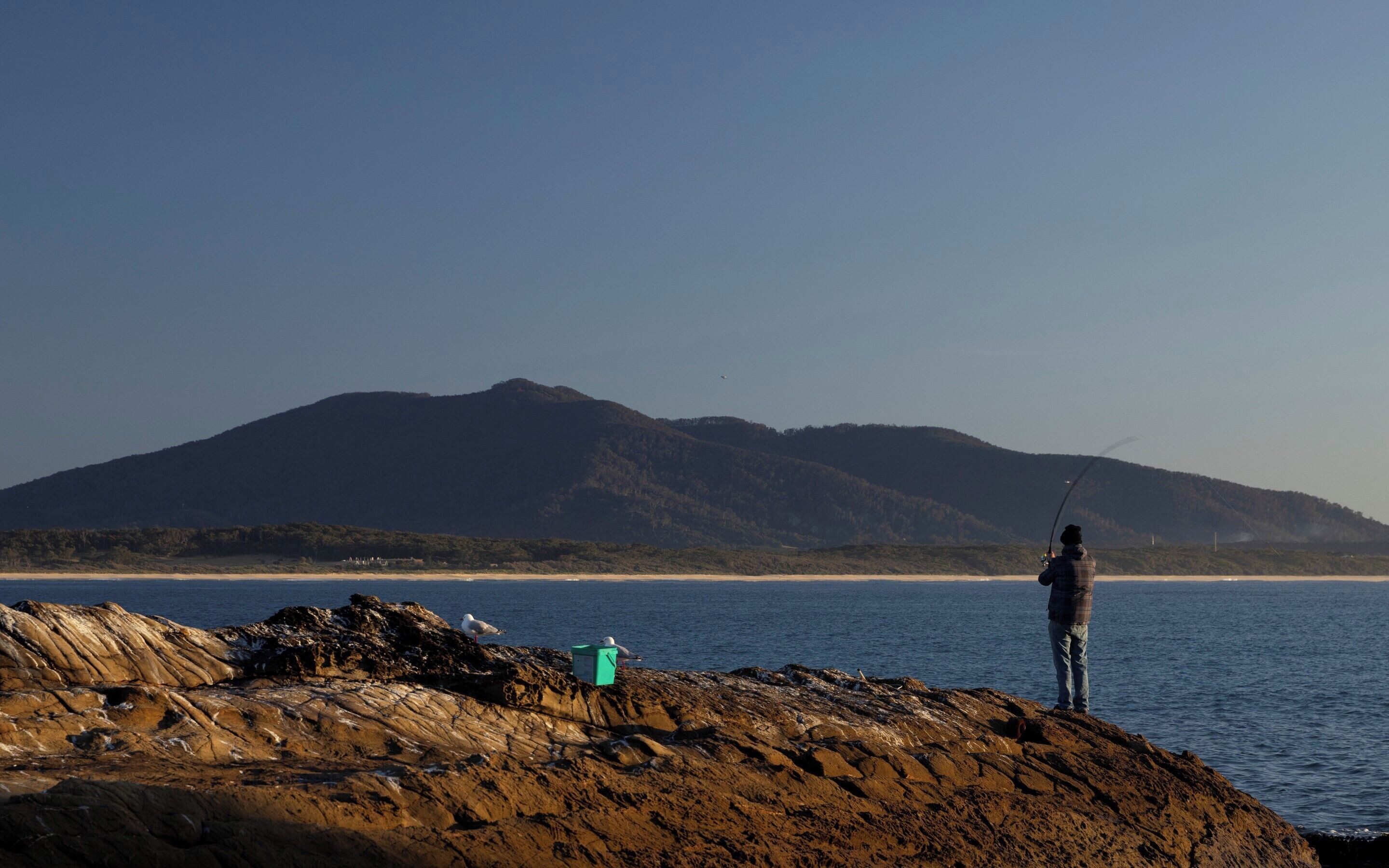Una persona pescando junto al mar, con una montaña al fondo.