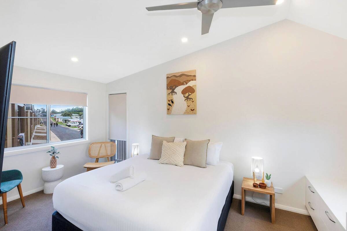 A modern hotel room with a large bed, a ceiling fan, a window with a city view, and a small bedside table with a lamp.