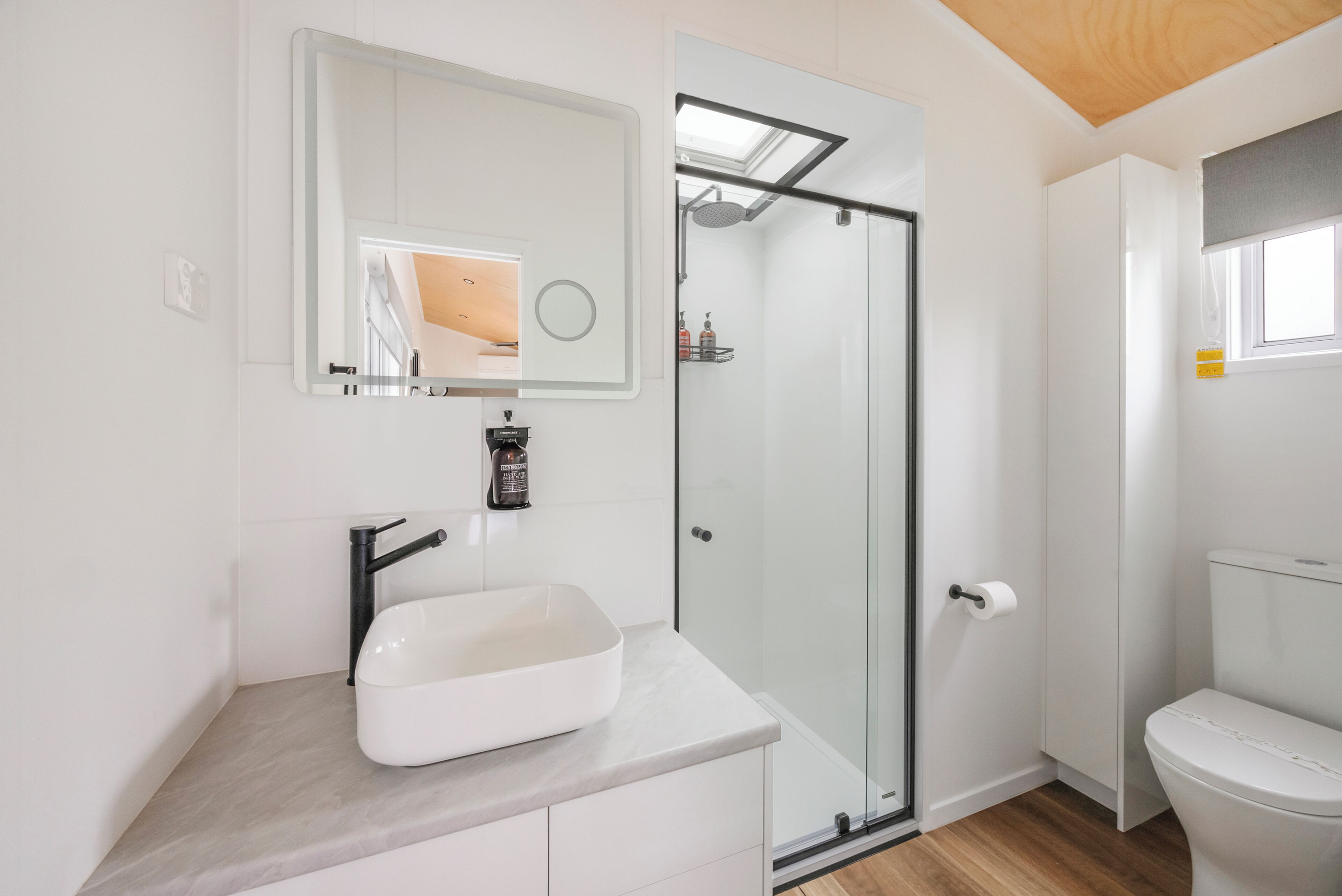 Et moderne badeværelse med glasvæg omkring brusebad, en hvid vask og toilet.
