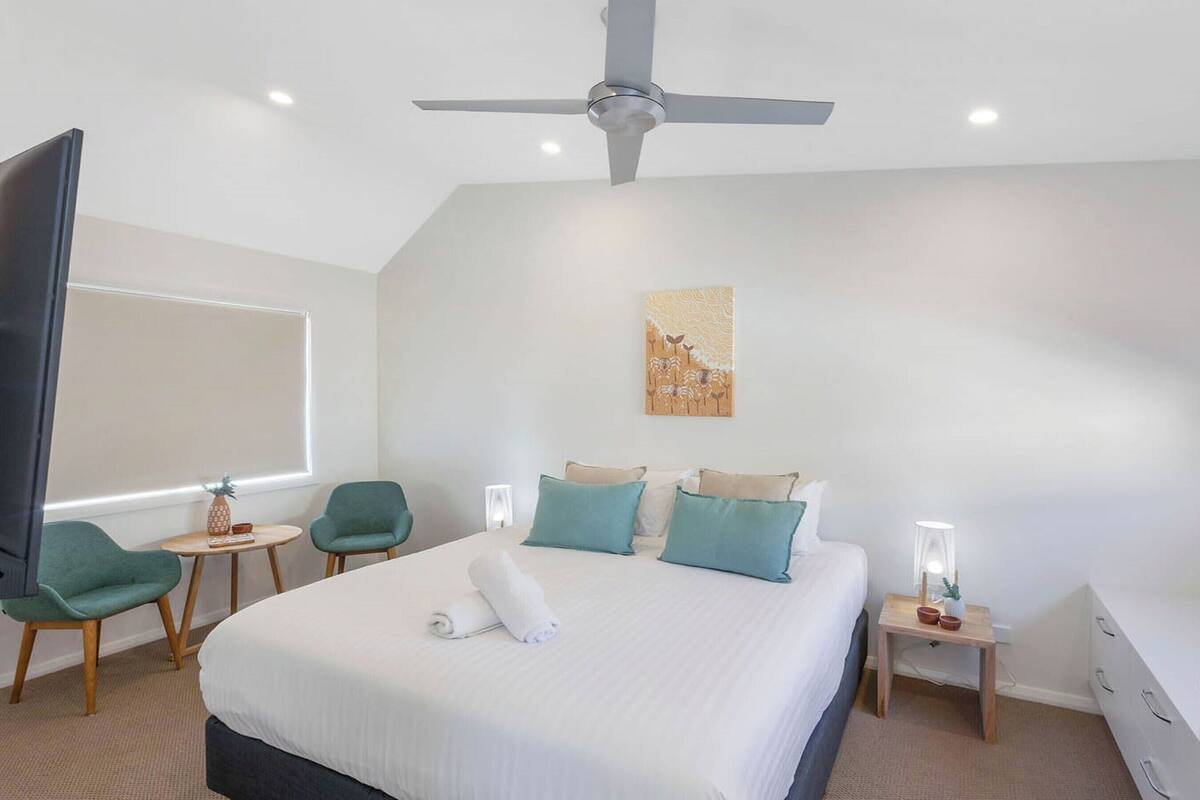 A modern hotel room with a large bed, a ceiling fan, a small desk with a lamp, a chair, and a wall-mounted artwork.