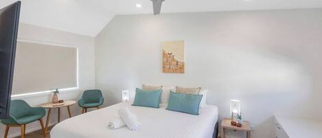 A modern hotel room with a large bed, a ceiling fan, a small desk with a lamp, a chair, and a wall-mounted artwork.