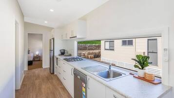 A modern kitchen with white cabinets, a dishwasher, a stove, and a sink. There is a window with a view of a stone wall and a plant on the counter.