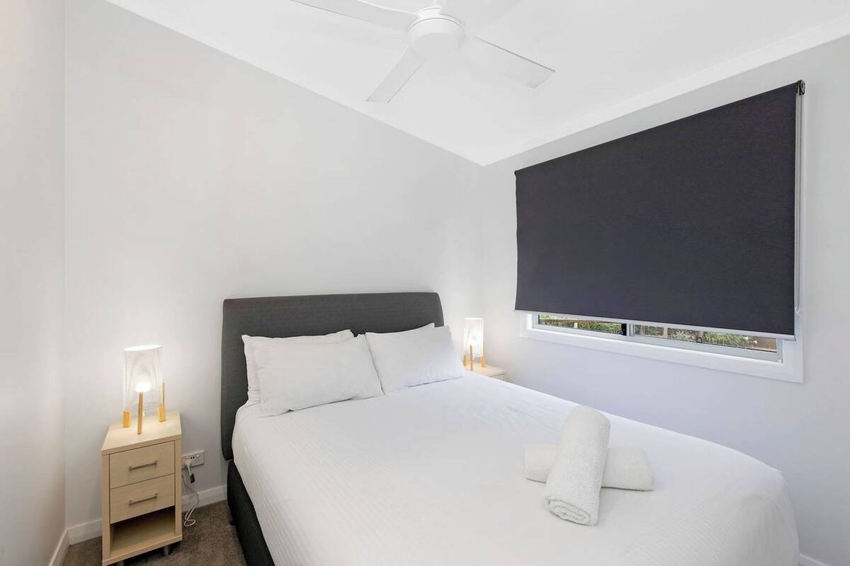 A neatly made bed with white linens, a bedside table with a lamp, and a ceiling fan.