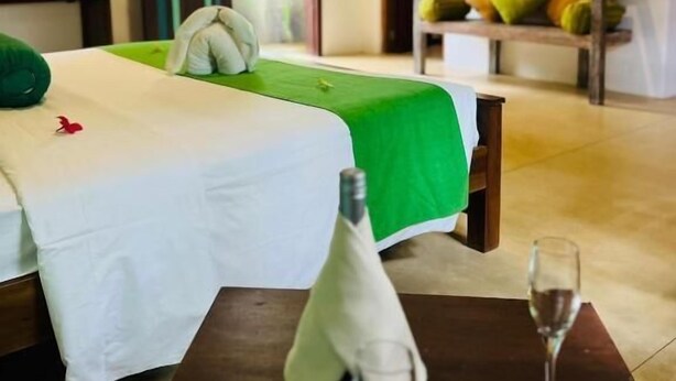 A hotel room with a bed, a green and white bedspread, a nightstand with a glass, and a television on the wall. - Garcinia Leaf (Hikkaduwa)