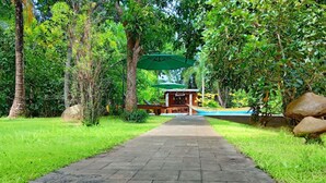 A pathway leading to a building with a green umbrella, surrounded by lush greenery and large rocks. - Garcinia Leaf (Hikkaduwa)