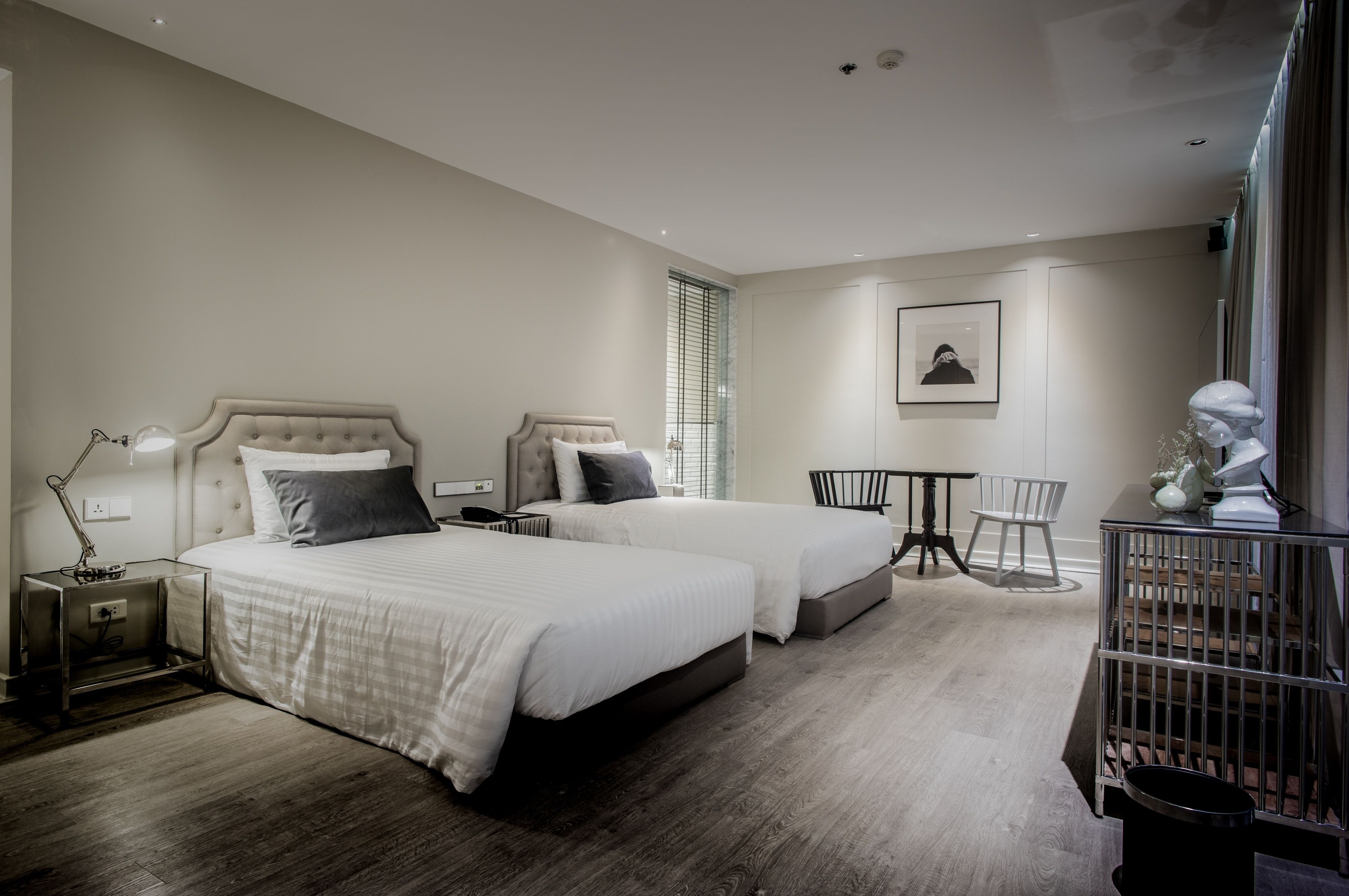 A hotel room with two beds, a bedside table, a chair, a small table, and a statue.