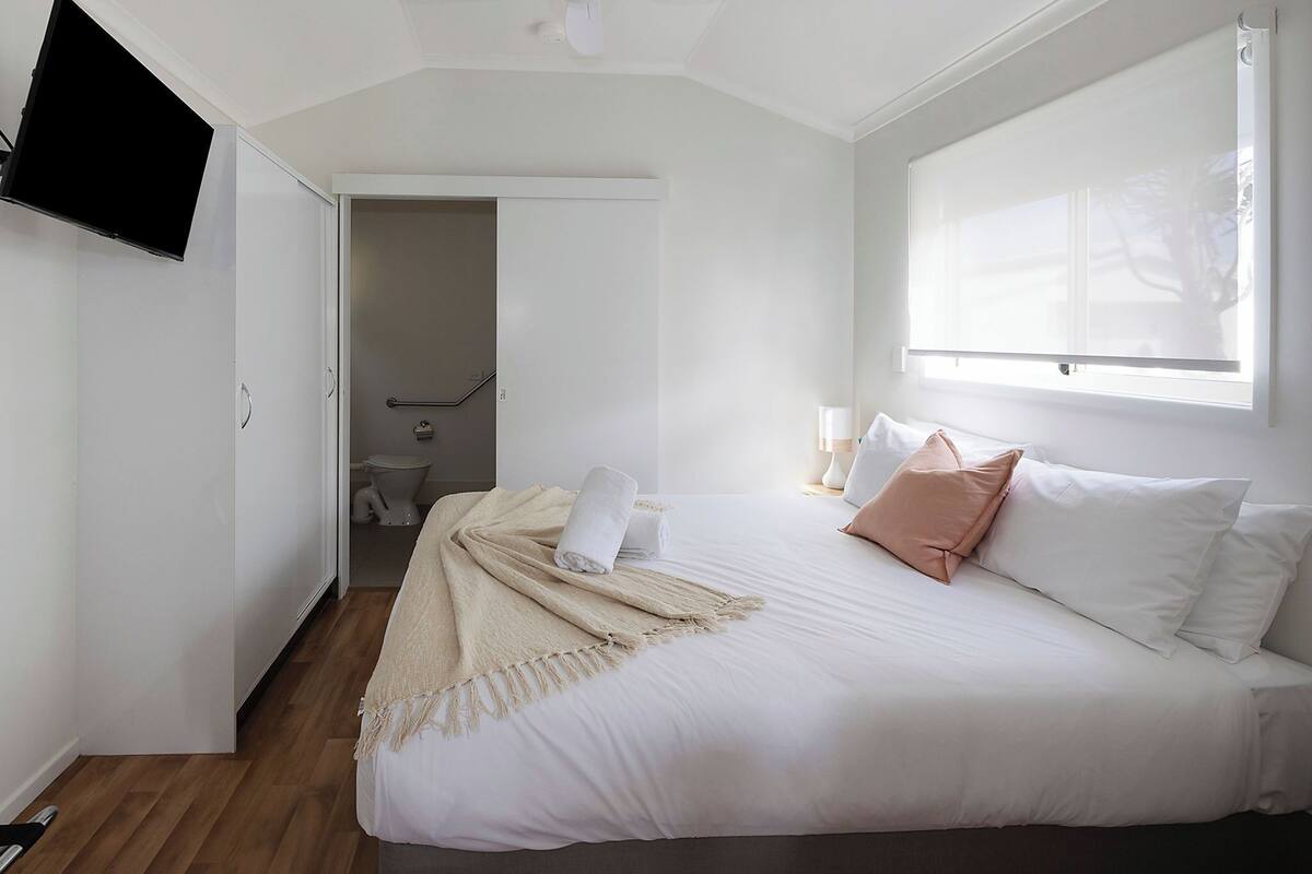 A bedroom with a large bed, a TV mounted on the wall, and a window with blinds.