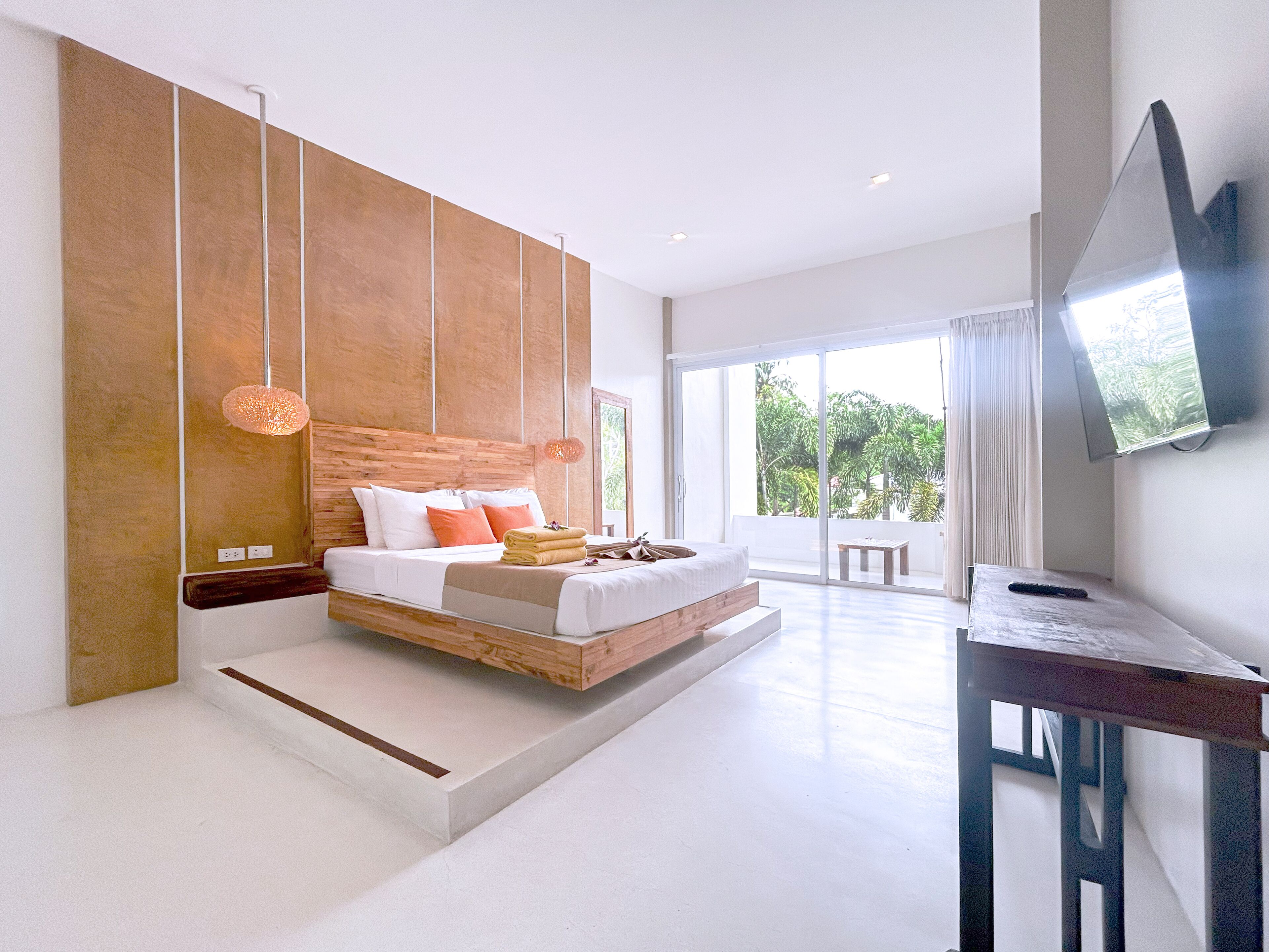 A modern bedroom with a large bed, a TV mounted on the wall, and a balcony with a view of greenery.
