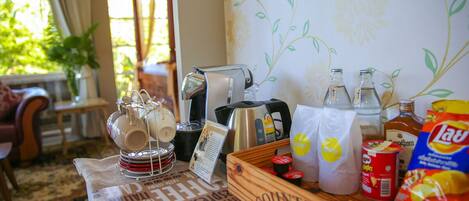 A table with coffee-making equipment, bags of coffee, and snacks in a cozy room with floral wallpaper and a view of greenery outside.