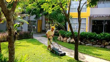 Un hombre corta el césped en un jardín cuidado, con un edificio amarillo al fondo.