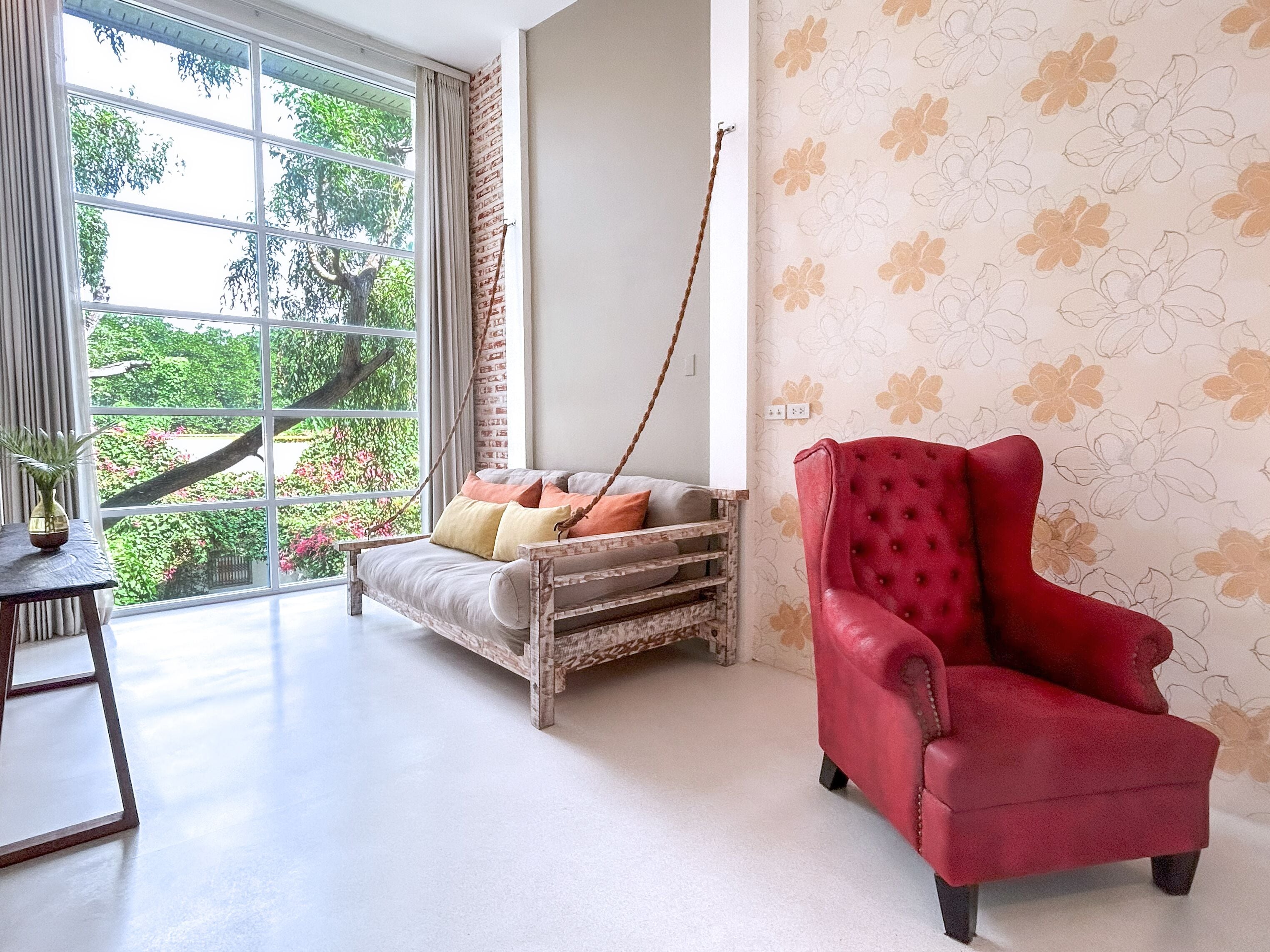 A room with a large window, a red armchair, a wicker sofa with cushions, and a wooden table with a vase.