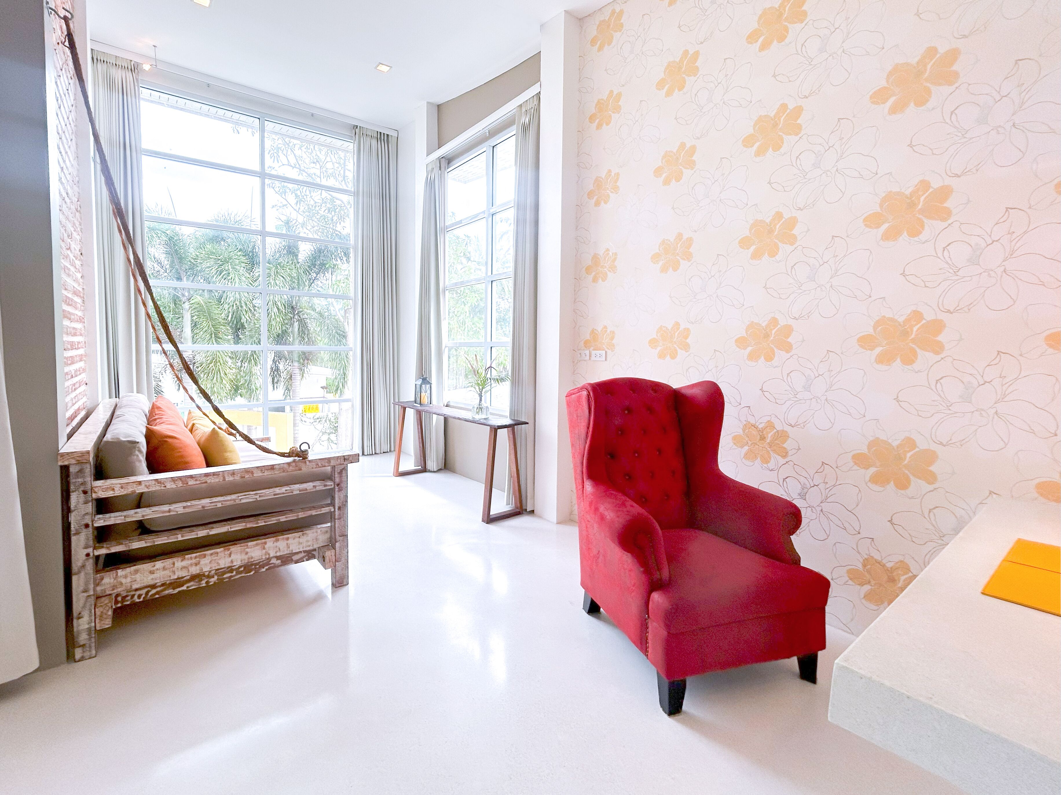 Una habitación con un sillón rojo, un banco de madera con cojines, ventanas amplias y un estampado floral en el papel tapiz.