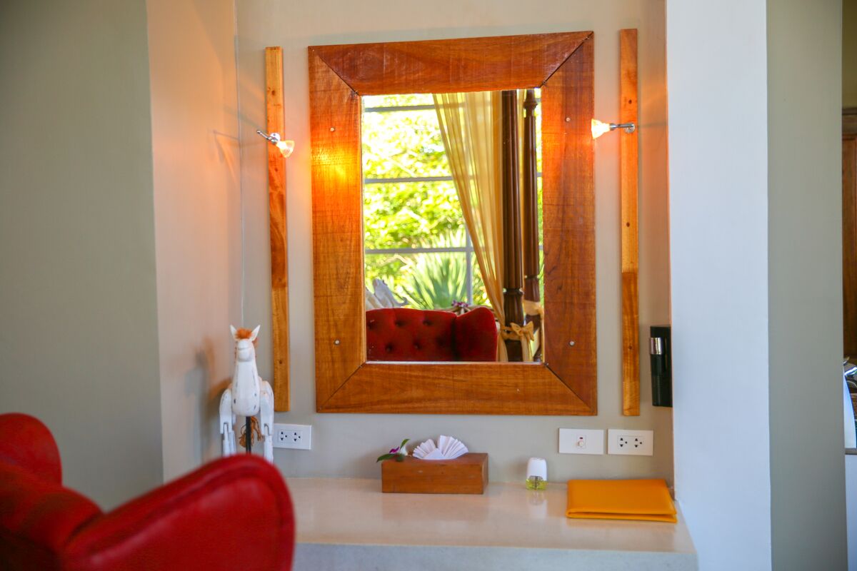 A bathroom with a wooden-framed mirror, a red chair, a white statue, and a small wooden box on the counter.