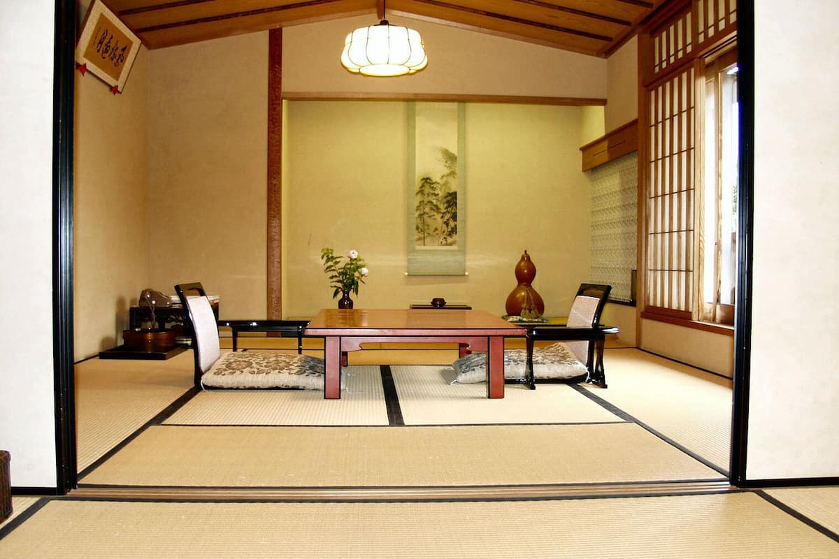 A tatami mat room with a low table, chairs, a hanging light fixture, a painting on the wall, and a window with vertical blinds.