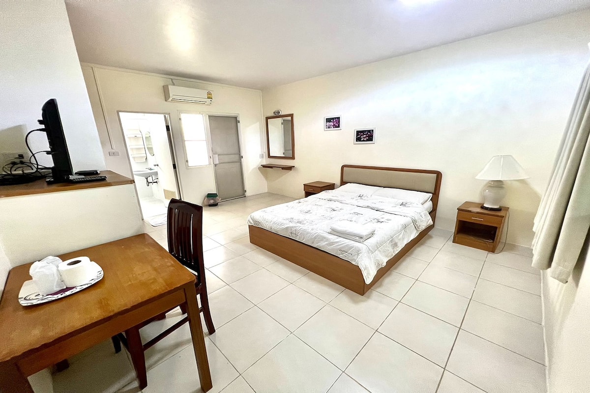 A hotel room with a bed, a desk with a computer, a chair, a bedside table with a lamp, and a bathroom visible through an open door.