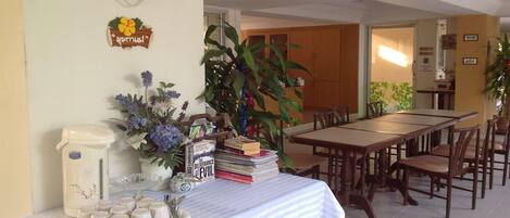 Un comedor con una mesa de madera larga y sillas, una estación de café con tazas y una cafetera, y una estantería con libros.