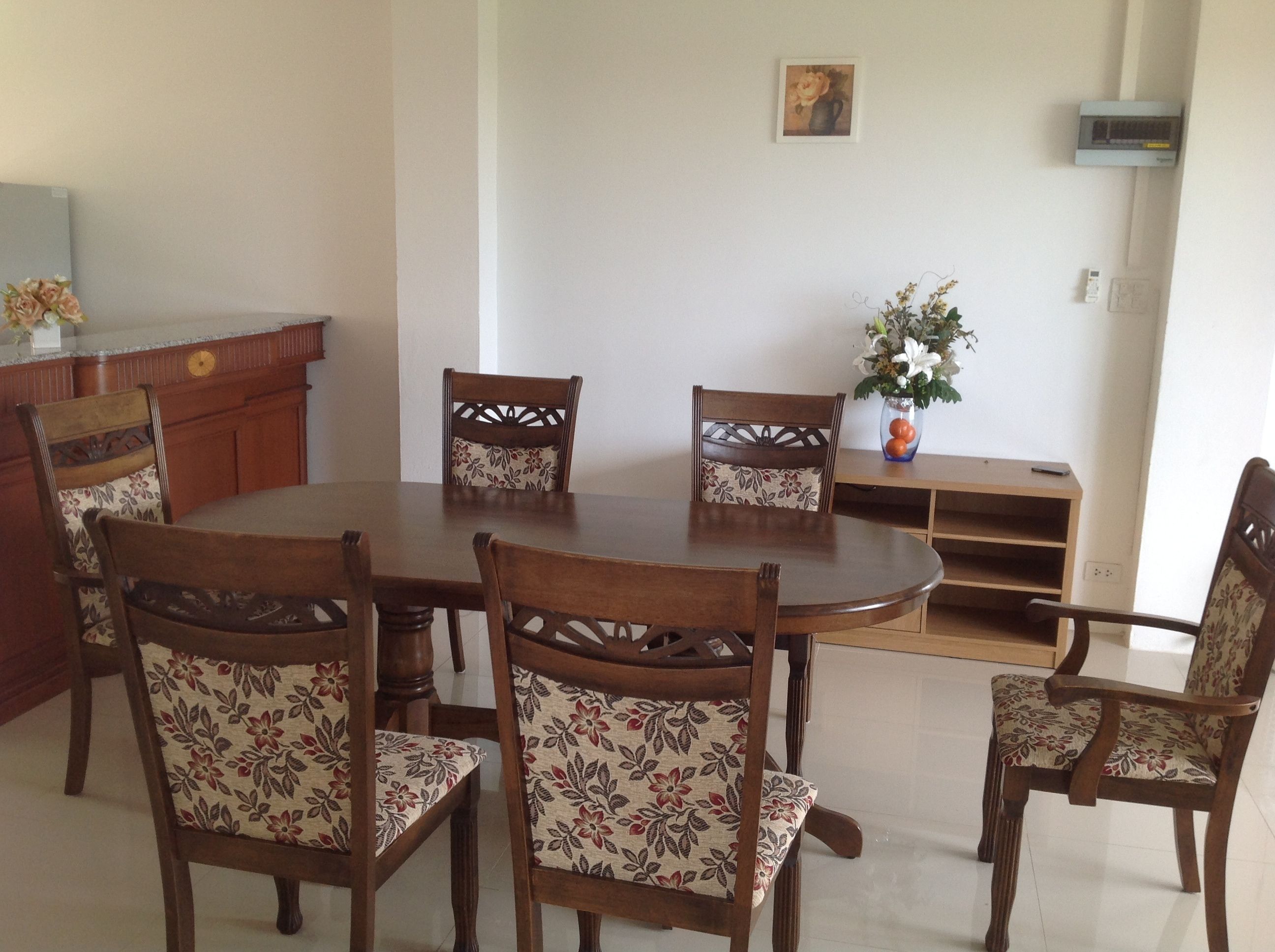 A dining area with a round wooden table surrounded by six chairs with floral upholstery. There is a wooden sideboard and a vase with flowers on a table to the right.