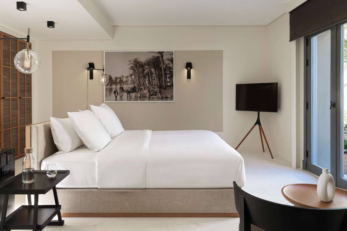 A modern bedroom with a large bed, a desk, a chair, a TV, and a window with a view.