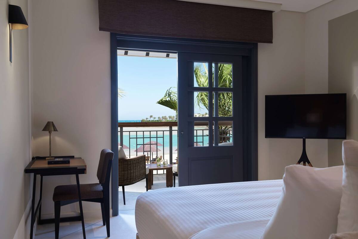 A hotel room with a bed, a desk with a lamp, a TV, and a balcony with a view of the beach.