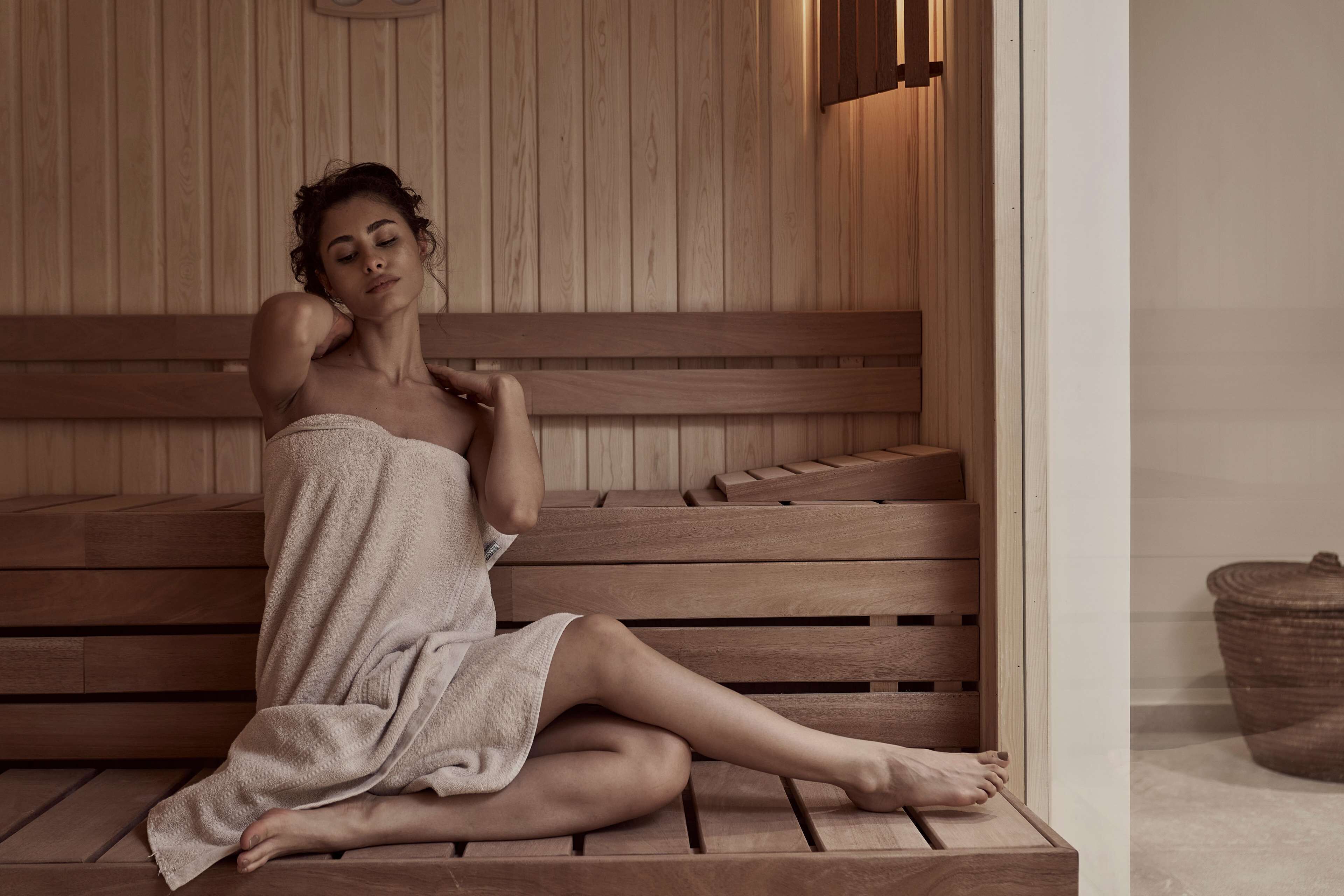 A person sitting on a wooden bench in a sauna room with wooden walls and a small trash bin in the corner.