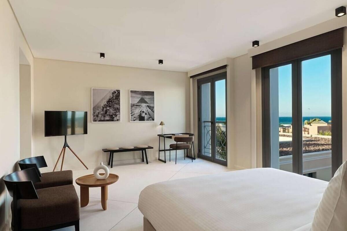A modern hotel room with a large bed, a desk, a chair, a TV, and a balcony with a view of the sea.