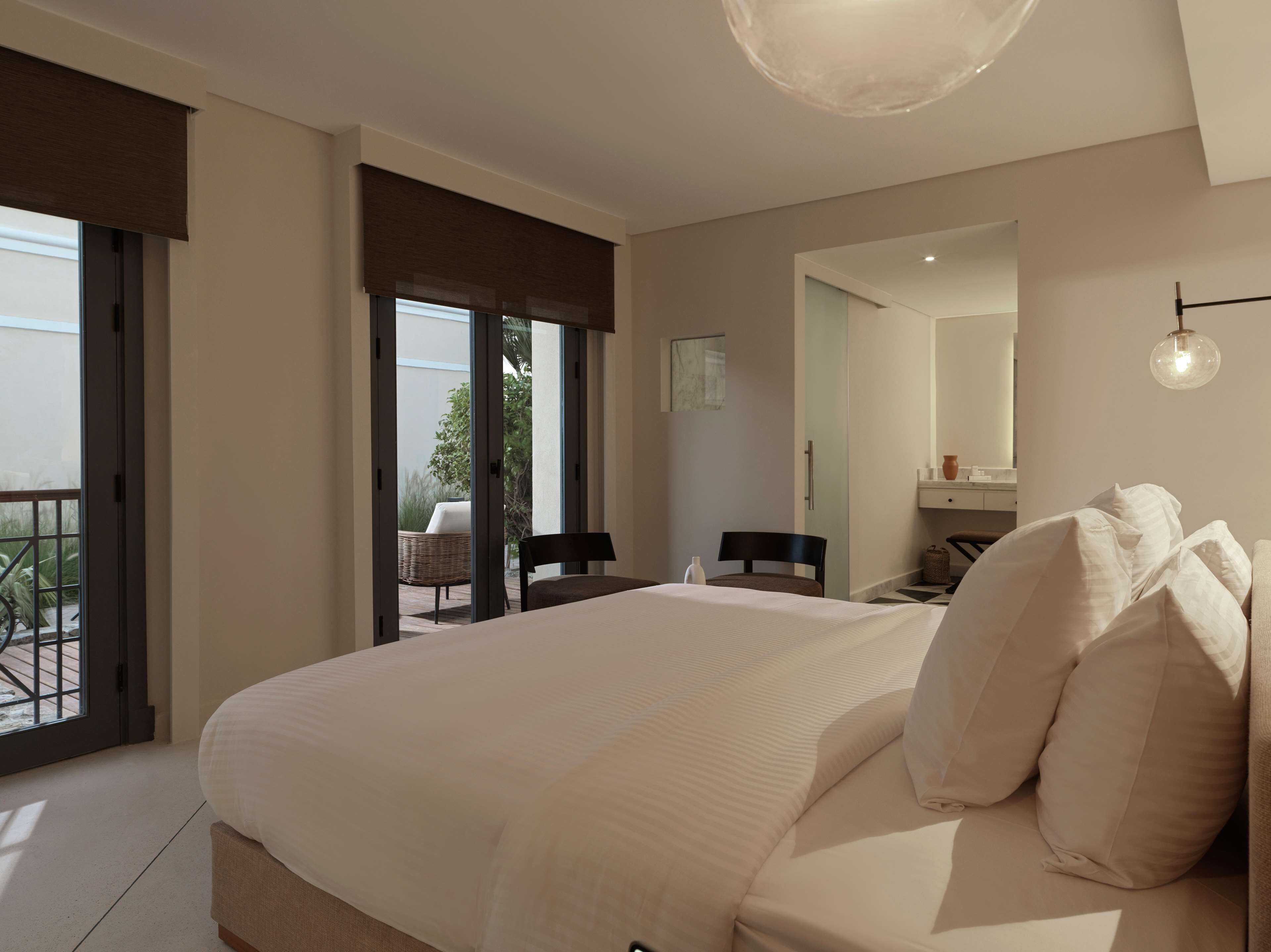 A bedroom with a large bed, a chair, a small table, a mirror, and a balcony with outdoor furniture.