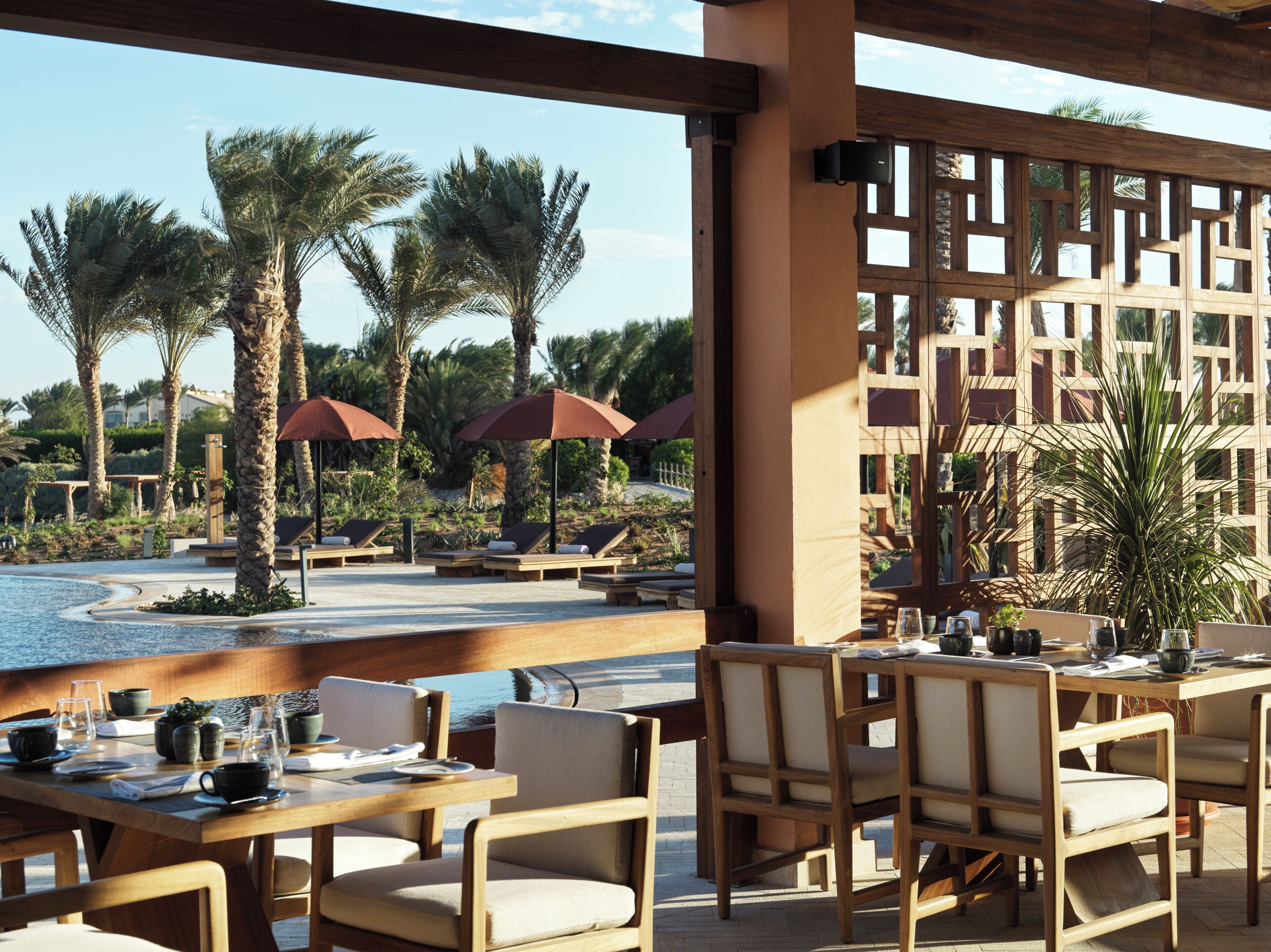 A resort pool area with lounge chairs and umbrellas, surrounded by palm trees and a view of a building in the background.