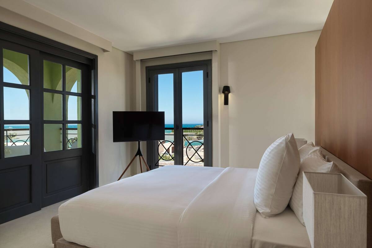 A hotel room with a large bed, a flat-screen TV, a balcony with a view of the sea, and a wooden headboard.