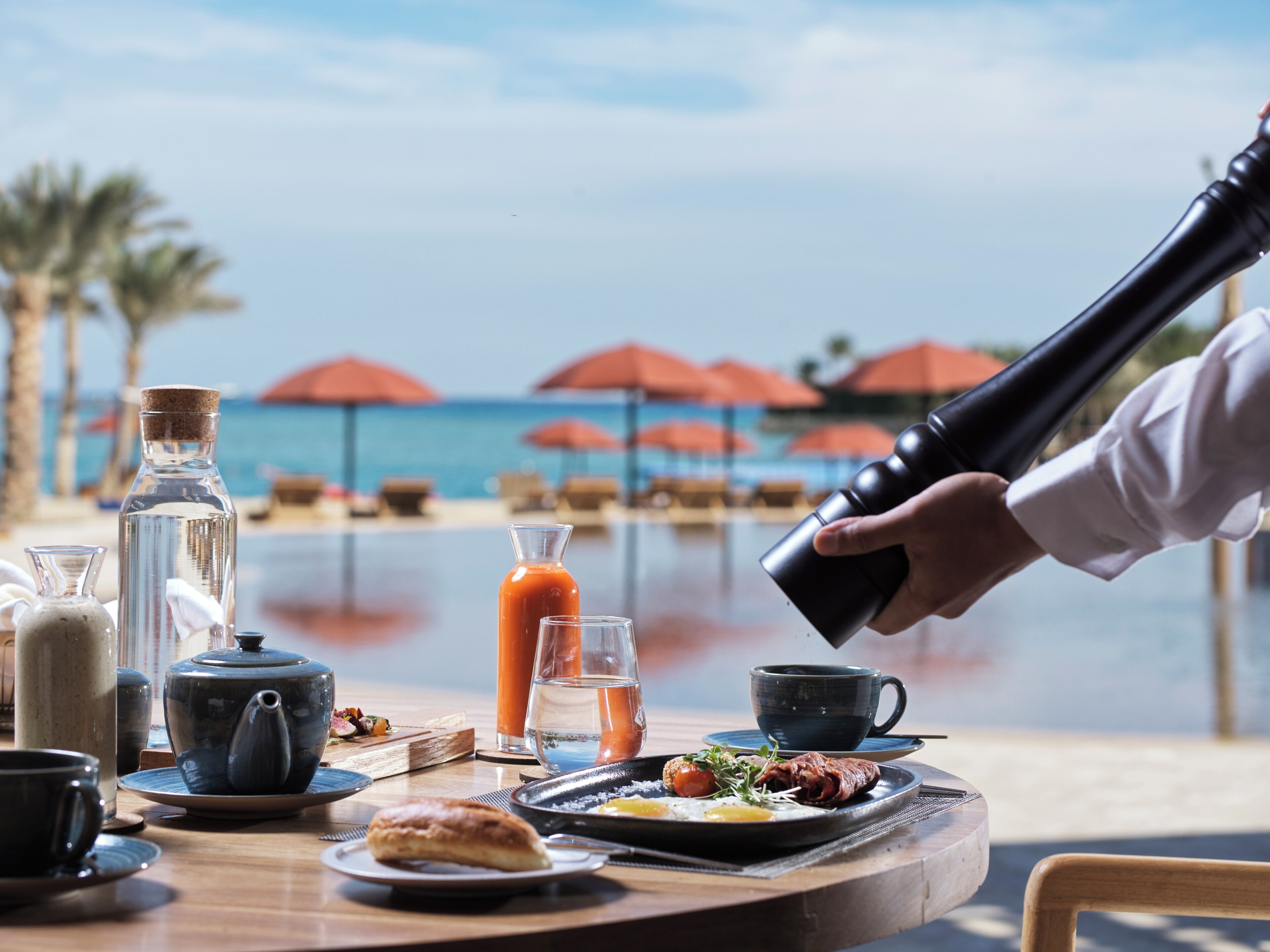 Una mesa de desayuno con diversos platos, incluyendo huevos, tostadas y un pastel, frente a una piscina y sombrillas.